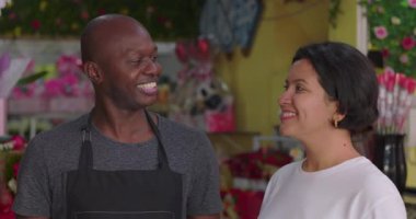 Çiçekçi ve müşteri birlikte gülümsüyor buketler, hediye süslemeleri ve canlı perakende vitrinleriyle çevrili neşeli bir adam ve kadın.