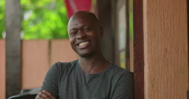 Afrika kökenli bir adam, açık havada duvara yaslanırken kollarını kavuşturarak güvenle gülümsüyor, şehir ortamında rahat bir ruh hali içinde.