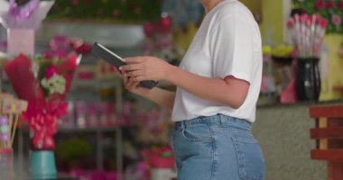 Çiçekçi dükkanındaki envanteri yönetmek için tablet kullanan kadın, buketler ve çiçeklerle çevrili dijital kayıtları kontrol eden girişimci, küçük işletme sahibi.