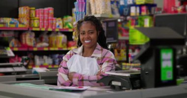 Kasada çalışan bir kadın markette kendine güvenerek gülümsüyor. Etrafı renkli ürün rafları ve yazar kasalarla çevrili.