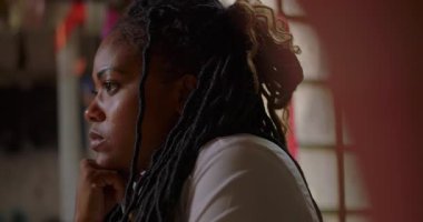 Derin düşüncelere dalmış, derin düşüncelere dalmış, ciddi bir ifadeyle ileri bakan, geniş pencereli, doğal ışık alan bir evde oturan Afro-Amerikan kadın.