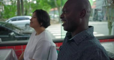 Gülümseyen Afro-Amerikalı erkek ve Latin kadın restoran masasında yan yana oturuyorlar. Pencereden sokak manzaralı neşeli bir ev sahnesi.