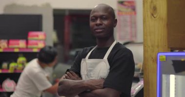 Afro-Amerikan şarküteri çalışanı arka planda Latin kadın müşteri alışverişiyle süpermarket koridorunda kesişiyor.