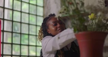 Gülümseyen Afrikalı Amerikalı kadın budama makasıyla çiçek düzenliyor, pencerenin yanında neşeyle çalışıyor, taze yeşil saplarla çiçek aranjmanı yapıyor.