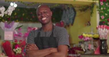 Çiçekçinin içinde kollarını kavuşturan ve gülümseyen, renkli çiçekler, buketler ve hediye süslemeleriyle çevrili gururlu küçük işletme sahibi.
