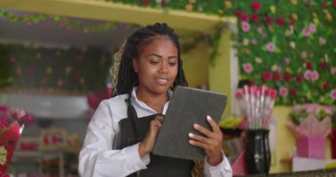 Afro-Amerikalı çiçekçi gülümserken tabletteki siparişleri inceliyor, renkli ve canlı çiçekçi dükkanında birden fazla iş yapıyor.