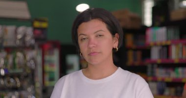 Ciddi bir Latin kadın süpermarket koridorunda duruyor, rengarenk günlük perakende ortamında doğal ışık ile odaklanmış ifade ediyor.