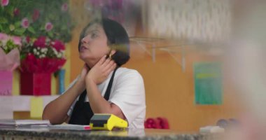 Çiçekçi dükkanında, gül buketleri, çiçek süslemeleri ve hediye ürünleriyle çevrili bir şekilde tezgaha yaslanmış bitkin bir çiçekçi.