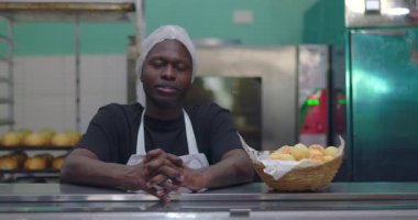 Kendine güvenen Afro-Amerikan fırın işçisi saç filesi ve önlükle tezgahın arkasında oturuyor ve profesyonel mutfak ortamında bir sepet ekmek rulosu ile oturuyor.