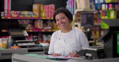 Süpermarket çalışanı kasanın arkasında gülümsüyor. Renkli rafları ve neşeli atmosferi olan sıradan bir market ortamı.