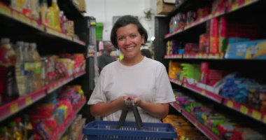 Gülümseyen Latin kadın marketin koridorunda alışveriş sepeti tutarken neşeli bir ifadeyle renkli atıştırmalık raflarının arasında duruyor.