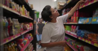 Market koridorunda abur cubur ve içeceklerle dolu alışveriş yaparken üst raftan bir şeyler almak için kollarını tamamen açan Latin bir kadın.