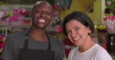 Çiçekçi ve müşteri çiçekçi dükkanında bir arada duruyorlar. Erkek ve kadın kameraya sıcak bir şekilde gülümsüyorlar. Etrafı buketlerle, hediyelerle ve canlı çiçeklerle çevrili.