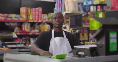 Kasanın arkasında duran süpermarket çalışanı, ciddi ifade, renkli kapalı market ortamı, günlük perakende atmosferi