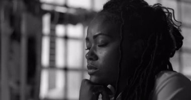 Düşünceli Afrikalı Amerikalı kadın, düşünceli bakıyor, çenesini dinlendiriyor, yoğun ifade, pencerenin yanında oturuyor, duygu ve odaklanma anı yakaladı.