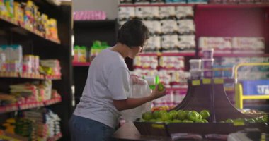 Süpermarket koridorunda taze yeşil elma toplayan Latin bir kadın paketlenmiş yiyecek rafları ve renkli ürünlerle çevrili.