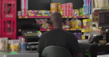 Afro-Amerikalı bir adam bakkal dükkanının arkasındaki kasada boş boş oturuyor. Müşteri bekliyor. Arka planda renkli ürünleri olan bir süpermarket.