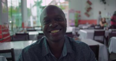 Restoranda sıcacık gülümseyen Afro-Amerikalı bir adam, rahat atmosferli sıradan bir resim, şehrin arka planında çekilmiş neşeli bir yüz ifadesi.