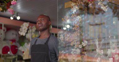 Yansıtıcı bir ifadeyle çiçekçinin yanında duran, önlük takan ve çiçekçinin penceresinden dışarı bakan Afro-Amerikalı bir adam.