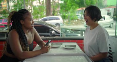 Restoran masasında iki farklı kadın konuşuyor ve gülüyor. Neşeli sohbetler, doğal ışık ve şehir manzaralı ev ortamları.