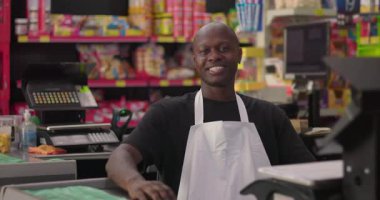 Süpermarket çalışanı kasada güvenle gülümsüyor. Renkli rafları ve rahat atmosferi olan sıradan bir market ortamında.