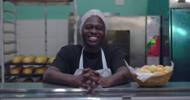 Önlüklü ve saç filesiyle sırıtan Afro-Amerikan bir adam fırın tezgahının arkasında duruyor. Elinde bir sepet ekmek ve ticari mutfakta rulo ekmek var.