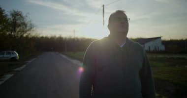 Gün batımında dışarıda yürüyen gözlüklü orta yaşlı bir adam, rahat ve düşünceli bir yüz ifadesi, kırsal alan, akşam ışığı, sakin atmosfer, kırsal huzur.