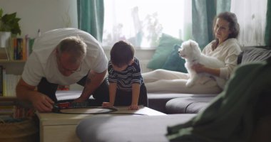 Aile, sıcak oturma odasında birlikte vakit geçirir. Büyükbaba, küçük çocukla oynar. Anne, evcil köpekle kanepede gülümser.