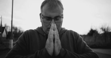 Elleri duada sıkışmış, başı eğik, derin düşüncelere dalmış, huzurlu kırsal alan, siyah beyaz, ruhani an, yansıma ve minnettarlık dolu bir adam.