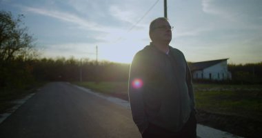 Orta yaşlı bir adam gün batımında kırsal bir yolda yürüyor, elleri ceplerinde, yansıtıcı ifade, huzurlu kırsal manzara, elektrik hatları, sakin alacakaranlık, kırsal ortam
