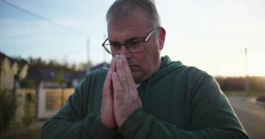 Dışarıda dua eden, elleri sımsıkı bağlı, aşağıya bakan, derin düşüncelere dalmış, günbatımında sakin kırsal bir sahne, minnettarlık ve maneviyat, huzurlu bir atmosfer.