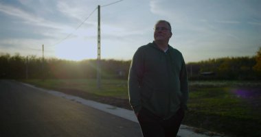 Orta yaşlı bir adam gün batımında kırsal bir yolda yürüyor, elleri ceplerinde, yansıtıcı ifade, huzurlu kırsal manzara, elektrik hatları, sakin alacakaranlık, kırsal ortam