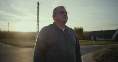 Karanlık çöktüğünde kırsal yolda duran bir adam, sakin bir ifadeyle, arka planda gün batımı parıltısı, kırsal bir manzara, huzurlu bir an, açık bir manzara, kırsal alanda yalnızlık.