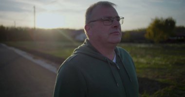 Gün batımında dışarıda yürüyen gözlüklü orta yaşlı bir adam, rahat ve düşünceli bir yüz ifadesi, kırsal alan, akşam ışığı, sakin atmosfer, kırsal huzur.