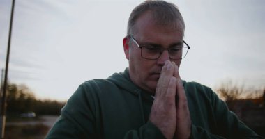 Elleri dua, yan görüş, yansıtıcı ve barışçıl ifade, sakin kırsal ortam, gün batımında ruhani an, şükran ve umut içinde kapalı bir adam.