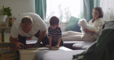 Aile, sıcak oturma odasında birlikte vakit geçirir. Büyükbaba, küçük çocukla oynar. Anne, evcil köpekle kanepede gülümser.