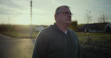 Karanlık çöktüğünde kırsal yolda duran bir adam, sakin bir ifadeyle, arka planda gün batımı parıltısı, kırsal bir manzara, huzurlu bir an, açık bir manzara, kırsal alanda yalnızlık.