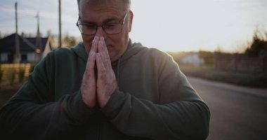 Elleri dua, yan görüş, yansıtıcı ve barışçıl ifade, sakin kırsal ortam, gün batımında ruhani an, şükran ve umut içinde kapalı bir adam.
