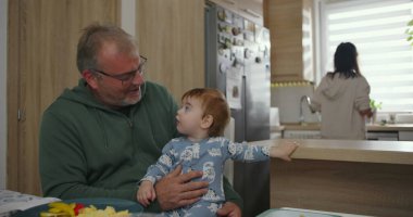 Baba mutfak tezgahında kucağında bebek, arka planda anne yemek hazırlıyor, sıcak aile anı, sabah rutini, birliktelik ve aşk bir arada.