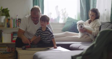 Aile, sıcak oturma odasında birlikte vakit geçirir. Büyükbaba, küçük çocukla oynar. Anne, evcil köpekle kanepede gülümser.