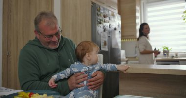 Baba mutfak tezgahında kucağında bebek, arka planda anne yemek hazırlıyor, sıcak aile anı, sabah rutini, birliktelik ve aşk bir arada.