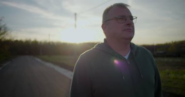 Gün batımında dışarıda yürüyen gözlüklü orta yaşlı bir adam, rahat ve düşünceli bir yüz ifadesi, kırsal alan, akşam ışığı, sakin atmosfer, kırsal huzur.