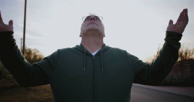 Dışarıdaki adam kollarını havaya kaldırmış, yukarıya bakıyor, minnettarlığını ve umudunu ifade ediyor, kırsal alanda ruhani bir an, sakin ve sakin, günbatımı atmosferi.