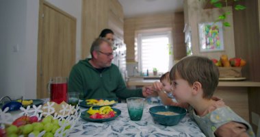 Baba karnını doyuran çocuk, aile yemeği sırasında yüksek sandalyede oturuyor, anne arka planda gülümsüyor, genç çocuk masada kaseyle oturuyor, sıcak aile etkileşimi içinde.