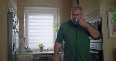 Mutfaktaki mavi bardaktan içen adam, sabah rutini, sıcak içecekten hoşlanan sıradan bir ev hayatı, rahat bir an, dekorlu modern bir mutfak ortamı.