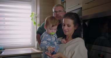 Anne bebeğini kucağında tutarken baba da ona bakıyor. Mutfak ortamında şefkatli ve ilgili bir aile etkileşimi yaratıyor.