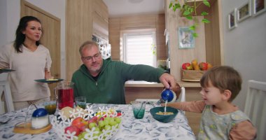 Baba yemek masasında çocuk için sütü kaseye koyuyor, anne gülümsüyor ve bebek bakıyor, ev mutfağında sıcak ve etkileşimli bir aile anı yaratıyor.