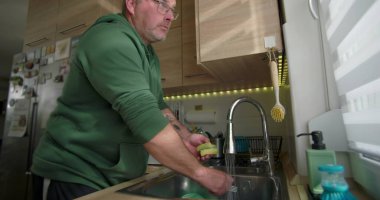 Adam mutfak lavabosunda bulaşık yıkıyor, akmakta olan bir kâseyi duruluyor, modern bir evde ev temizliği yapıyor.