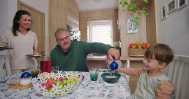 Baba yemek masasında çocuk için sütü kaseye koyuyor, anne gülümsüyor ve bebek bakıyor, ev mutfağında sıcak ve etkileşimli bir aile anı yaratıyor.