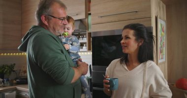 Anne elinde kahve fincanı tutarken, baba mutfakta bebeği tutarken, sakin bir aile etkileşimi ve günlük hayatta takım çalışması sergiliyor.